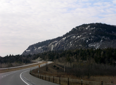 [The highway starts in the lower left heading toward the center then snakes back out of view to the left. Towering above it are tree-topped mountains dappled with snow.]